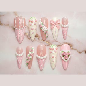 May include: A collection of ten almond-shaped artificial nails, each adorned with intricate designs. The nails feature a palette of pink, white, and cream colors, with embellishments like pearls, bows, cherries, and floral patterns. The designs vary, showcasing a range of decorative nail art.