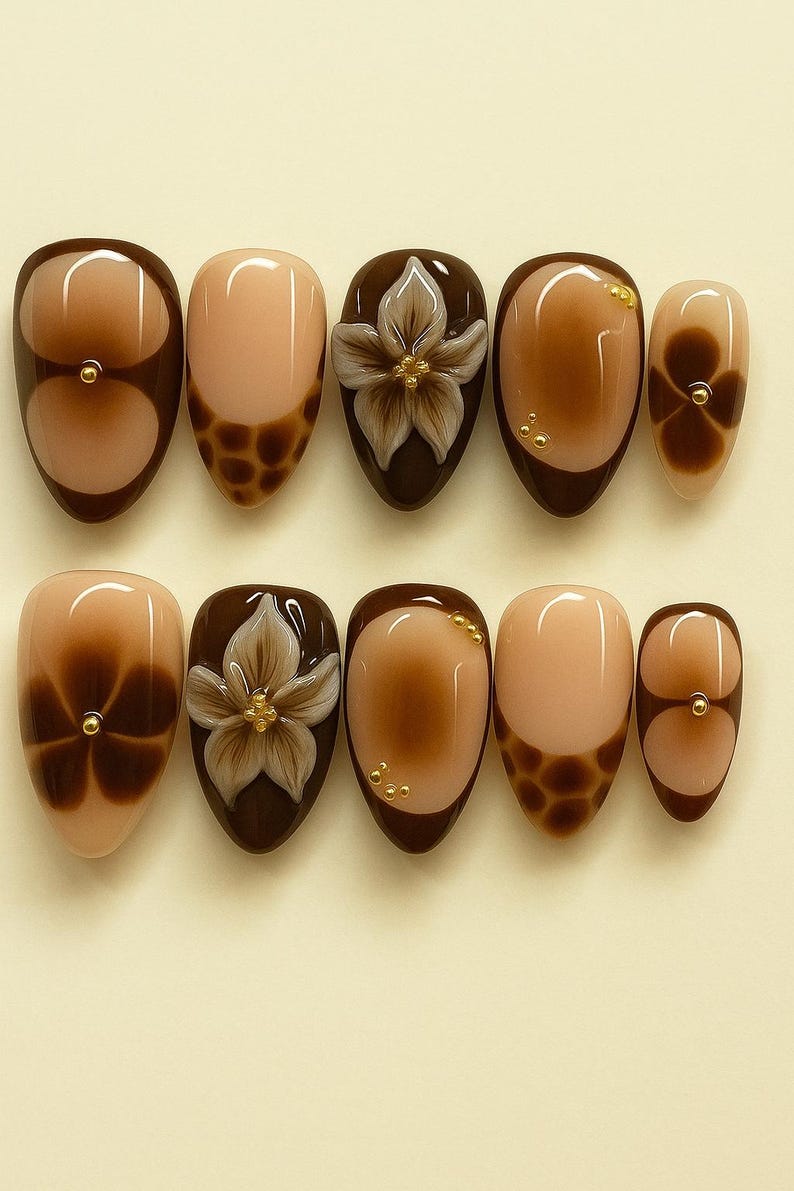 May include: A set of ten almond-shaped false nails. The nails feature a variety of designs, including a brown gradient, a floral design with gold accents, and a brown leopard print. The colour palette includes shades of brown and beige.