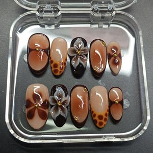 May include: A set of ten oval-shaped, press-on nails in a clear plastic case. The nails feature various designs, including brown floral patterns, a 3D white flower with gold accents, and a brown and tan gradient with gold dots. The nails are glossy.