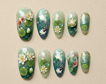 May include: A set of ten almond-shaped artificial nails with a water lily and koi fish design. The nails feature a gradient of green and blue hues, with 3D floral and fish accents. The design includes water lilies, lily pads, and koi fish.