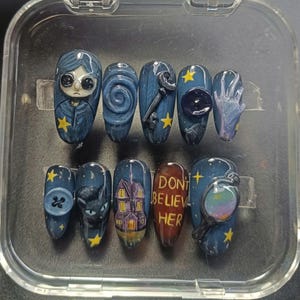May include: A set of ten custom press-on nails in dark blue and grey. Designs include a character, a key, a button, a cat, a house, and the text "DON'T BELIEVE HER". The nails are decorated with yellow stars.