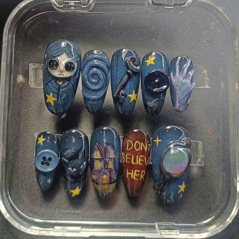 May include: A set of ten custom press-on nails in dark blue and grey. Designs include a character, a key, a button, a cat, a house, and the text "DON'T BELIEVE HER". The nails are decorated with yellow stars.