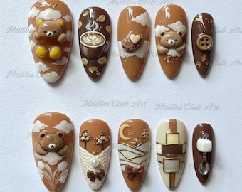 Op de afbeelding: Een set van tien amandelvormige kunstnagels met diverse ontwerpen. De nagels hebben bruine, beige en witte kleuren met 3D-elementen zoals teddyberen, koffiekopjes, wolken en strikken. De tekst "Malibu Club Art" is zichtbaar.
