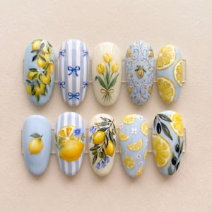Pode incluir: Um conjunto de dez unhas artificiais pintadas com vários desenhos. Os desenhos incluem limões, fatias de limão, laços e padrões florais em tons de amarelo, azul e branco. As unhas são ovais e dispostas em fila.