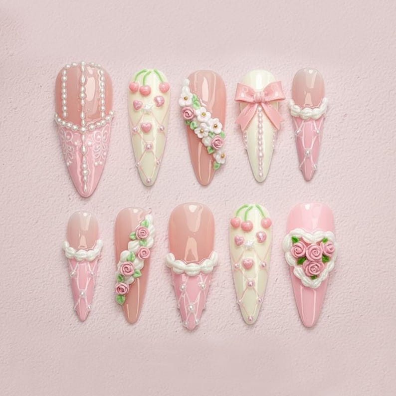May include: A collection of ten almond-shaped artificial nails in various designs. The nails feature pink, white, and cream colors, adorned with pearls, bows, floral accents, and heart-shaped decorations. The background is a soft pink.
