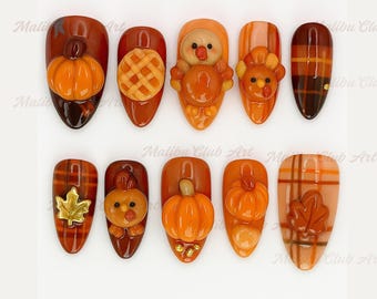 May include: A set of ten fall-themed nail art designs. Each nail features a different design, including pumpkins, turkeys, and plaid patterns in shades of orange, brown, and gold. The nails are almond-shaped.