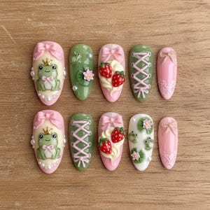 May include: A set of ten press-on nails with various designs. The nails feature frog, strawberry, and lace patterns, with pink, green, and white color schemes. Some nails have bows and pearl accents, while others have a lace-up design.