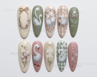 Può includere: Una collezione di dieci unghie finte ovali, ognuna decorata con intricati disegni dipinti a mano. I disegni presentano cigni, fiori, fiocchi e altri elementi decorativi in morbidi colori pastello, tra cui rosa, verde e beige.