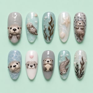 Op de afbeelding: Een set van tien ovale kunstnagels met gedetailleerde zeeotterontwerpen. De nagels bevatten verschillende aquatische elementen, waaronder zeeotters, schelpen, parels en zeewier, in grijstinten, blauw en wit. De nagels staan tegen een lichtgroene achtergrond.