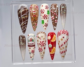 May include: A set of ten almond-shaped, festive nail designs. The nails feature various holiday-themed decorations, including gingerbread men, candy canes, and snow globes. Colors include red, white, brown, and gold, with glitter accents. "Malibu Club Art" is visible.