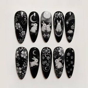 May include: Black and white nail art featuring various designs. The designs include rabbits, moons, flowers, and stars. The nails are almond-shaped and have a glossy finish.