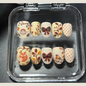 May include: A set of ten press-on nails with an autumn theme. The nails feature designs such as a fox, coffee art, bows, leaves, and birds. The base color is off-white, with accents in orange, brown, and red. The nails are in a clear plastic case.