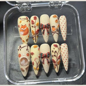 May include: A set of ten artificial nails with a fall theme. The nails are off-white with various designs, including a fox, leaves, a bow, a bird, and polka dots. The nails are in a clear plastic case.