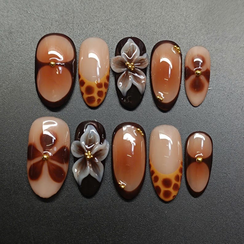 May include: A collection of ten artificial nails in diverse designs. The nails showcase a blend of brown, beige, and white hues. Some nails feature a floral design, while others have a gradient or animal print pattern. Gold accents are present on several nails.