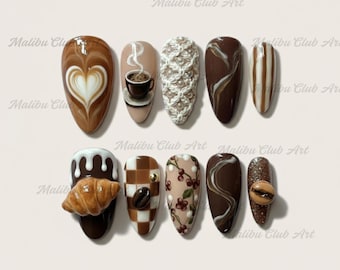 May include: A collection of ten almond-shaped artificial nails with coffee-themed designs. The nails feature various designs, including latte art, coffee cups, and a croissant. Colors include brown, beige, white, and gold. The text "Malibu Club Art" is visible.