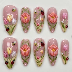 May include: A set of ten almond-shaped false nails. Each nail is painted a pale pink with white polka dots. The nails are decorated with 3D tulips, ladybirds, and bees. Some nails feature vines.