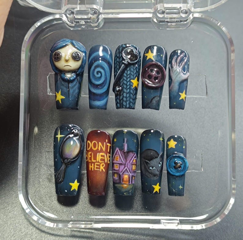 May include: A set of ten press-on nails with a dark blue base, featuring various designs from a movie. Designs include a character, a spiral, a key, a button, a hand, a mirror, the text "DON'T BELIEVE HER", a house, a cat, and a button. Yellow stars are scattered throughout.