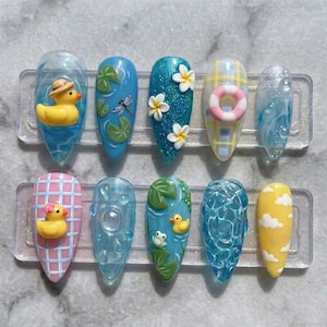 May include: A set of ten artificial nails with summer-themed designs. The nails feature various designs, including a yellow rubber duck, water lilies, a dragonfly, flowers, a life preserver, and clouds. The nails are in shades of blue, yellow, and pink.
