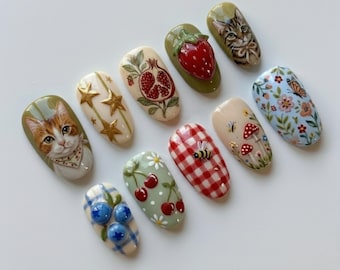 Könnte beinhalten: Zehn künstliche Fingernägel mit verschiedenen Designs. Die Designs umfassen eine Katze, Sterne, einen Granatapfel, eine Erdbeere, einen Schmetterling, Blumen, Pilze, eine Biene, Kirschen und Blaubeeren. Die Nägel sind in verschiedenen Farben bemalt, darunter Rot, Grün, Blau und Weiß.