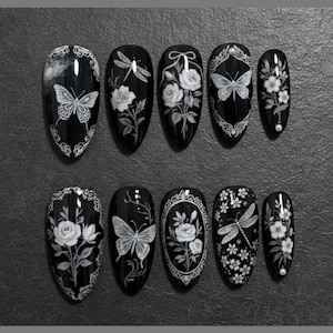 May include: A set of ten black artificial nails with white floral and insect designs. The designs include butterflies, dragonflies, and roses. Each nail has a glossy finish and a different design. The nails are arranged on a gray surface.