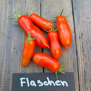 Puede incluir: Un conjunto de tomates Flaschen de color rojo brillante con tallos verdes, dispuestos sobre una superficie de madera rústica. Un pequeño letrero negro con la palabra "Flaschen" en escritura blanca se coloca cerca de los tomates. Los tomates son alargados y lisos.