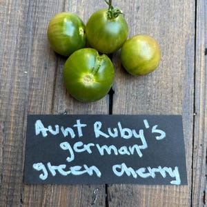 Könnte beinhalten: Vier grüne Cherrytomaten auf einer Holzoberfläche. Die Tomaten variieren in Grüntönen und Gelb. Ein schwarzes Schild mit weißem Text lautet "Aunt Ruby's german green cherry".