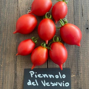 May include: A collection of bright red Piennolo del Vesuvio tomatoes with green stems, arranged on a rustic wooden surface. The tomatoes are oval-shaped with a pointed end. A black sign with white text reads "Piennolo del Vesuvio".