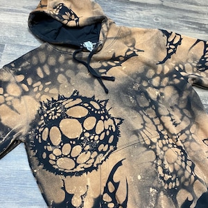 May include: A black hooded sweatshirt featuring a bleached, tan-colored abstract pattern across the fabric. The design includes a large, circular, spiky graphic on the front, a front kangaroo pocket, and black drawstring cords at the neckline.