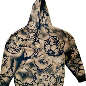 May include: A beige hoodie with a unique pattern of dark blue, circular, and web-like designs. The hoodie features a front pocket, a drawstring hood, and black cuffs. The overall design gives the hoodie a distinctive, artistic appearance.