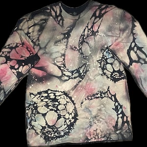 May include: Long-sleeved shirt with a unique tie-dye pattern. The shirt features a blend of colors, including pink, black, and gray, with intricate, organic-shaped designs. The sleeves and neckline are solid black. This shirt is a statement piece.