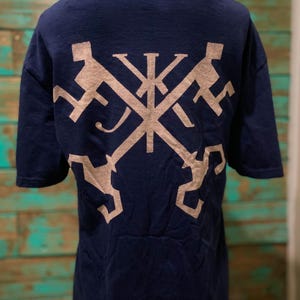 Op de afbeelding: Marineblauw T-shirt met een tan grafisch ontwerp op de rug. Het ontwerp is een gestileerd kruis met geometrische vormen. Het shirt heeft korte mouwen en een ronde hals. De achtergrond is een turquoise en bruin houten oppervlak.