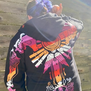 Puede incluir: Una sudadera negra con capucha que presenta un gran gráfico colorido en la espalda y la manga. El diseño incorpora un patrón radial en tonos rosa, naranja y blanco, con intrincadas líneas de estilo mecánico y formas geométricas.