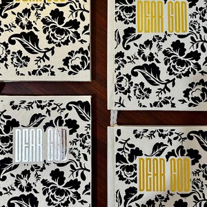 Puede incluir: Cuatro diarios rectangulares con cubierta de tela color crema y un patrón floral de damasco negro. Cada diario tiene el texto "DEAR GOD" impreso en una fuente vertical y negrita, tres con lámina dorada y uno con lámina plateada.