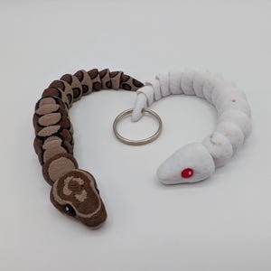 Op de afbeelding: Een handgemaakte slangen sleutelhanger, met een tweekleurig ontwerp. De ene helft is bruin en beige, de andere is wit. De slangenkop heeft zwarte ogen en een gedetailleerd patroon. De witte kop heeft rode ogen. Een zilveren sleutelring verbindt de twee helften.