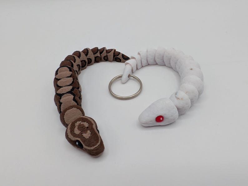 Op de afbeelding: Een handgemaakte slangen sleutelhanger, met een tweekleurig ontwerp. De ene helft is bruin en beige, de andere is wit. De slangenkop heeft zwarte ogen en een gedetailleerd patroon. De witte kop heeft rode ogen. Een zilveren sleutelring verbindt de twee helften.