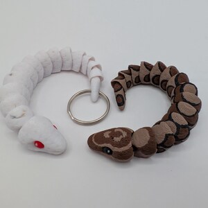May include: Two snake-shaped keychains. One is white with red eyes, the other is brown and tan with black markings. Both are coiled and connected to a silver key ring. The keychains are likely made of fabric.