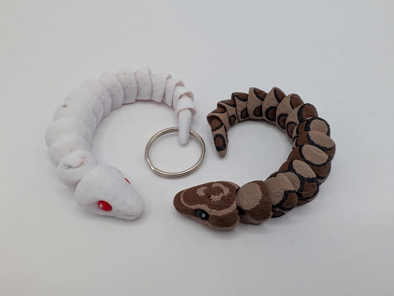 Op de afbeelding: Twee slangen-vormige sleutelhangers. E&eacute;n is wit met rode ogen, de andere is bruin en bruin met zwarte markeringen. Beide zijn opgerold en verbonden met een zilveren sleutelring. De sleutelhangers zijn waarschijnlijk gemaakt van stof.