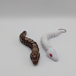 Op de afbeelding: Twee slangvormige beeldjes. E&eacute;n is bruin en beige, met een gedetailleerd patroon. De andere is wit met rode ogen en een sleutelhanger. Beide staan op een witte ondergrond. De slangenbeeldjes zijn waarschijnlijk speelgoed of decoratieve items.