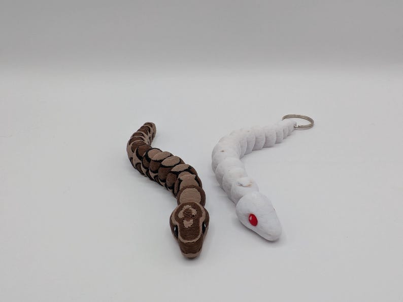 Op de afbeelding: Twee slangvormige beeldjes. E&eacute;n is bruin en beige, met een gedetailleerd patroon. De andere is wit met rode ogen en een sleutelhanger. Beide staan op een witte ondergrond. De slangenbeeldjes zijn waarschijnlijk speelgoed of decoratieve items.