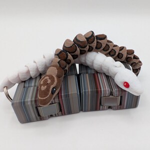 Op de afbeelding: Een 3D-geprint gearticuleerd slangen speelgoed in wit, bruin en bruin. De slang rust op twee rechthoekige blokken met een gestreept patroon. De ogen van de slang zijn rood en hij heeft een sleutelhanger.