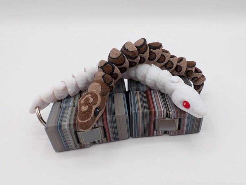 Op de afbeelding: Een 3D-geprint gearticuleerd slangen speelgoed in wit, bruin en bruin. De slang rust op twee rechthoekige blokken met een gestreept patroon. De ogen van de slang zijn rood en hij heeft een sleutelhanger.