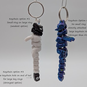 Op de afbeelding: Twee sleutelhangers met verschillende bevestigingsopties. De ene is wit en zwart, de andere blauw en paars. Tekst geeft de sterkte van elke optie aan: kleine ring aan grote ring, geen kleine ring en groot sleutelhangergat.