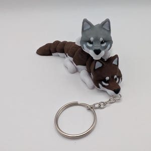 May include: A handmade keychain featuring two wolf figurines. The top wolf is gray, and the bottom wolf is brown, both with white accents. The keychain has a silver-colored metal ring and chain.