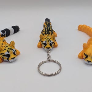 May include: Three cheetah figurines, one is a keychain with a silver ring. The cheetah figurines are orange and black with white spots. The cheetah figurines are in a variety of poses.