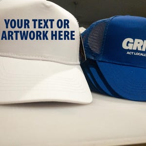 May include: A white trucker hat with the blue text YOUR TEXT OR ARTWORK HERE and a blue trucker hat with the white text GRNDED ACT LOCALLY, THINK GLOBALLY. Both hats feature mesh back panels and solid front panels with curved brims.