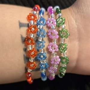 Könnte beinhalten: Fünf Perlenarmbänder mit blütenförmigen Perlen in Orange, Blau, Lila und Grün. Die Armbänder sind auf dem Handgelenk einer Person übereinander gelegt.