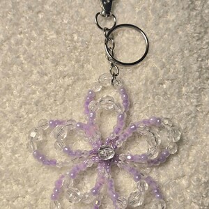 Könnte beinhalten: Ein Schlüsselanhänger in Blumenform mit klaren und lavendelfarbenen Perlen, einem silbernen Verschluss und Ring. In der Mitte der Blume befindet sich ein klarer Edelstein. Dekoratives Accessoire.