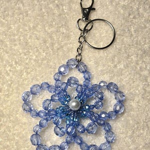 Könnte beinhalten: Ein blauer Schlüsselanhänger mit einer blumenförmigen Perlenverzierung und einem silbernen Verschluss und Ring. Die Blume besteht aus facettierten blauen Perlen mit einer Perle in der Mitte. Der Schlüsselanhänger ist für Schlüssel oder Taschen gedacht.