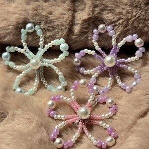 Könnte beinhalten: Drei Perlenblumen-Ornamente in Pastellfarben. Eine ist hellblau, eine andere rosa und lila, und die dritte ist lila. Jede Blume hat eine große Perle in der Mitte und ist vor einem verschwommenen, hellbraunen Hintergrund platziert.
