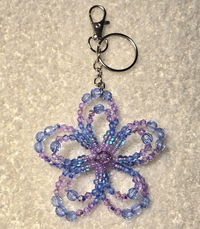 K&ouml;nnte beinhalten: Ein Schl&uuml;sselanh&auml;nger mit einer blumenf&ouml;rmigen Perlenarbeit in Blau- und Lilat&ouml;nen. Das Blumendesign besteht aus kleinen, facettierten Perlen, die zu einer f&uuml;nfbl&auml;ttrigen Form aufgef&auml;delt sind. Oben sind ein silberfarbener Karabiner und ein Schl&uuml;sselring befestigt.
